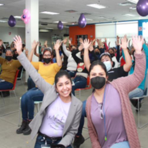 Two women, one with a mask, in front of a group of people with everyone raising one arm up and smiling