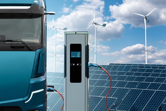 A truck charges an electric vehicle, with wind turbines visible in the background, symbolizing renewable energy use
