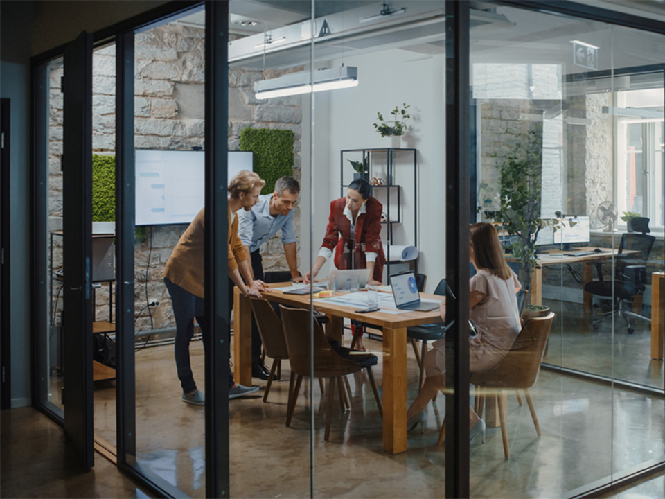 Diverse group of people gathered around a table in a glass-walled conference room, with several wireless devices