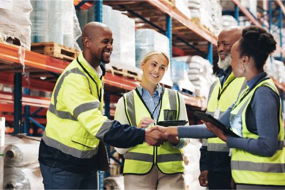 a diverse group of men and women in warehouse wearing safety vest greeting each other.